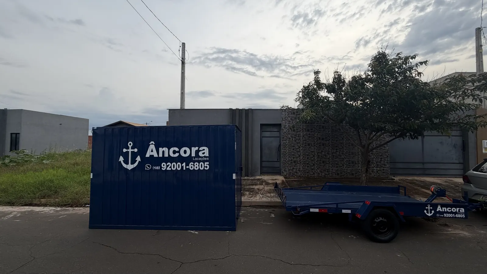 Container almoxarifado azul da Âncora Locações com o adesivo da marca e o telefone (18) 92001-6805, na rua em frente a uma obra, ao lado de uma carretinha azul da Âncora